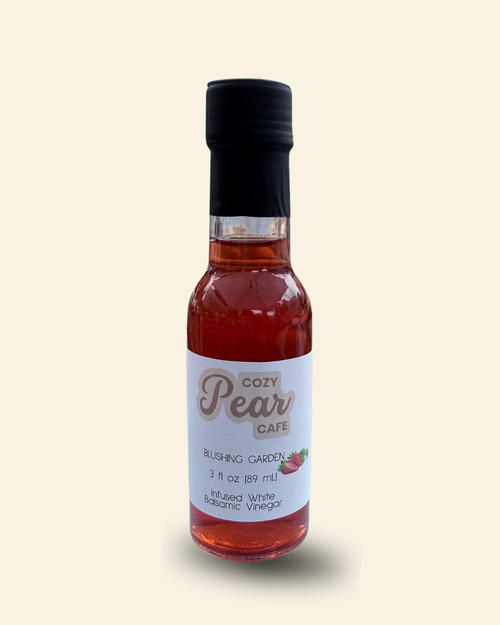 A small glass bottle of Cozy Pear Cafe Blushing Garden flavored white vinegar, filled with a reddish-amber liquid and topped with a black cap, set against a solid cream background.