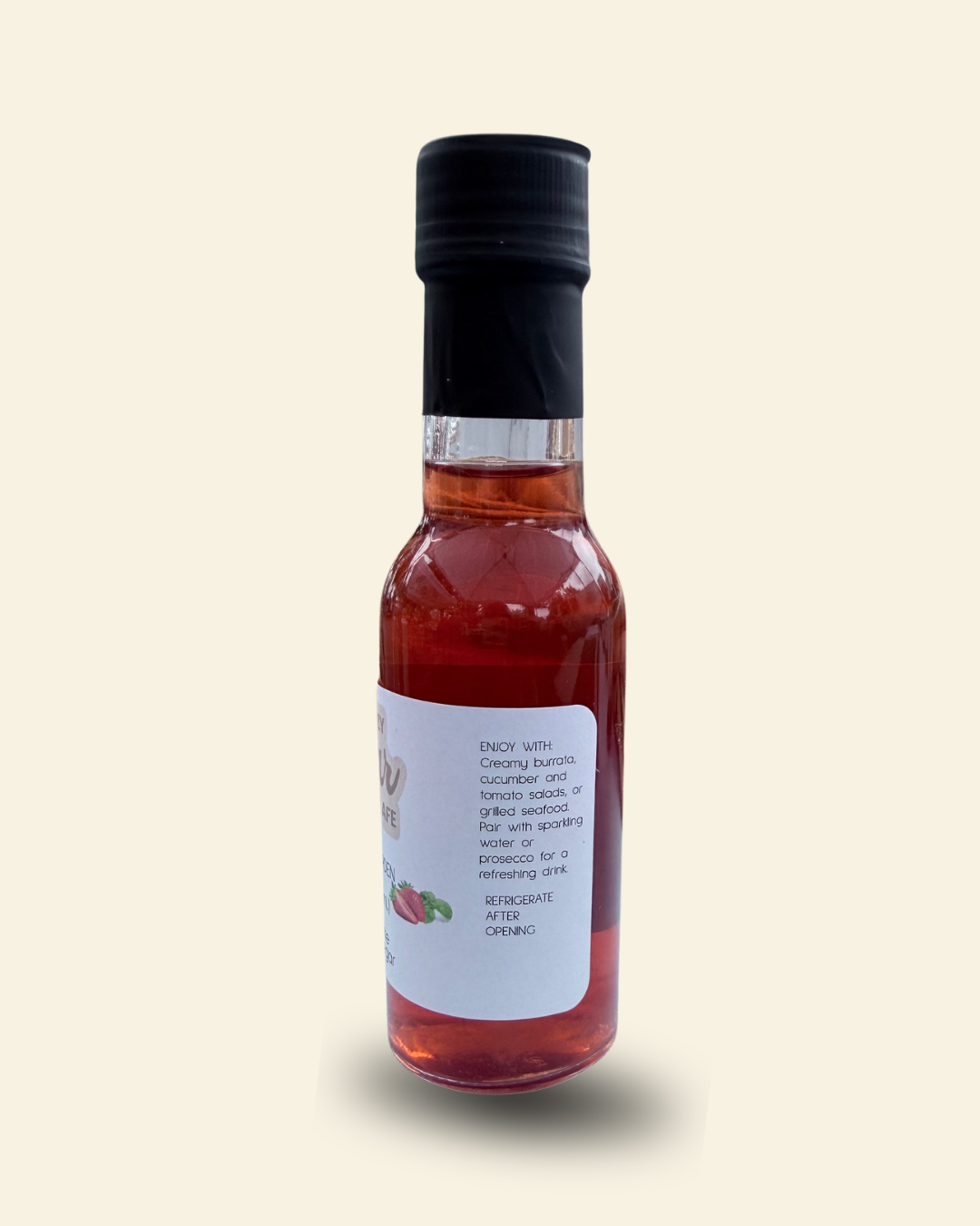 A small glass bottle of Cozy Pear Cafe Blushing Garden flavored white vinegar, filled with a reddish-amber liquid and topped with a black cap, set against a solid cream background.
