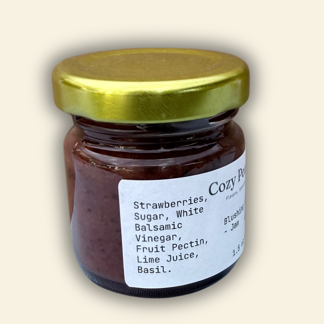 A jar of Cozy Pear Cafe Blushing Garden - Jam, featuring a white label and a metallic gold lid, set against a solid cream-colored background.