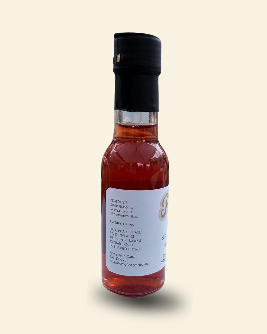 A small glass bottle of Cozy Pear Cafe Blushing Garden flavored white vinegar, filled with a reddish-amber liquid and topped with a black cap, set against a solid cream background.