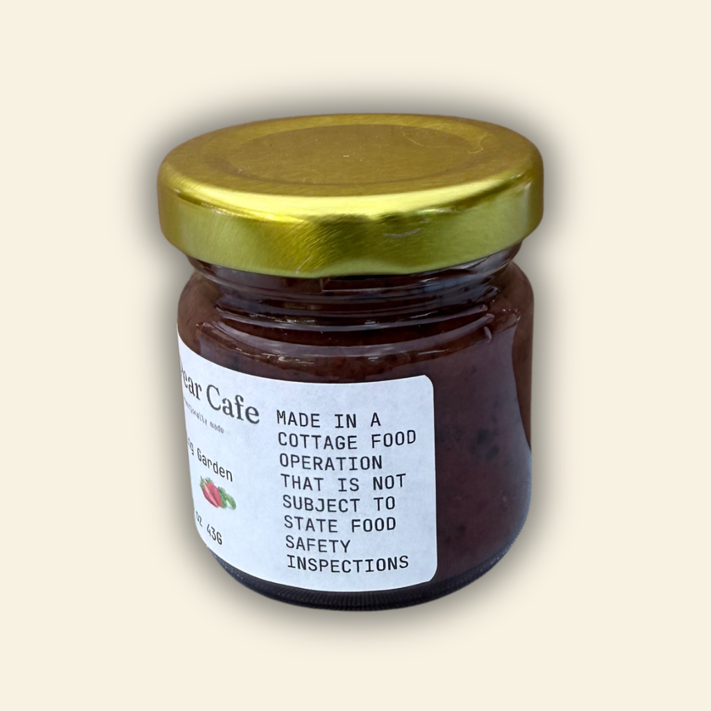 A jar of Cozy Pear Cafe Blushing Garden - Jam, featuring a white label and a metallic gold lid, set against a solid cream-colored background.