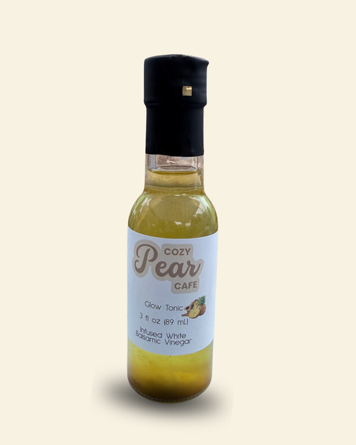 A bottle of Cozy Pear Cafe Glow Tonic, made from distilled white balsamic vinegar, featuring a white label and a black cap, set against a solid cream background.