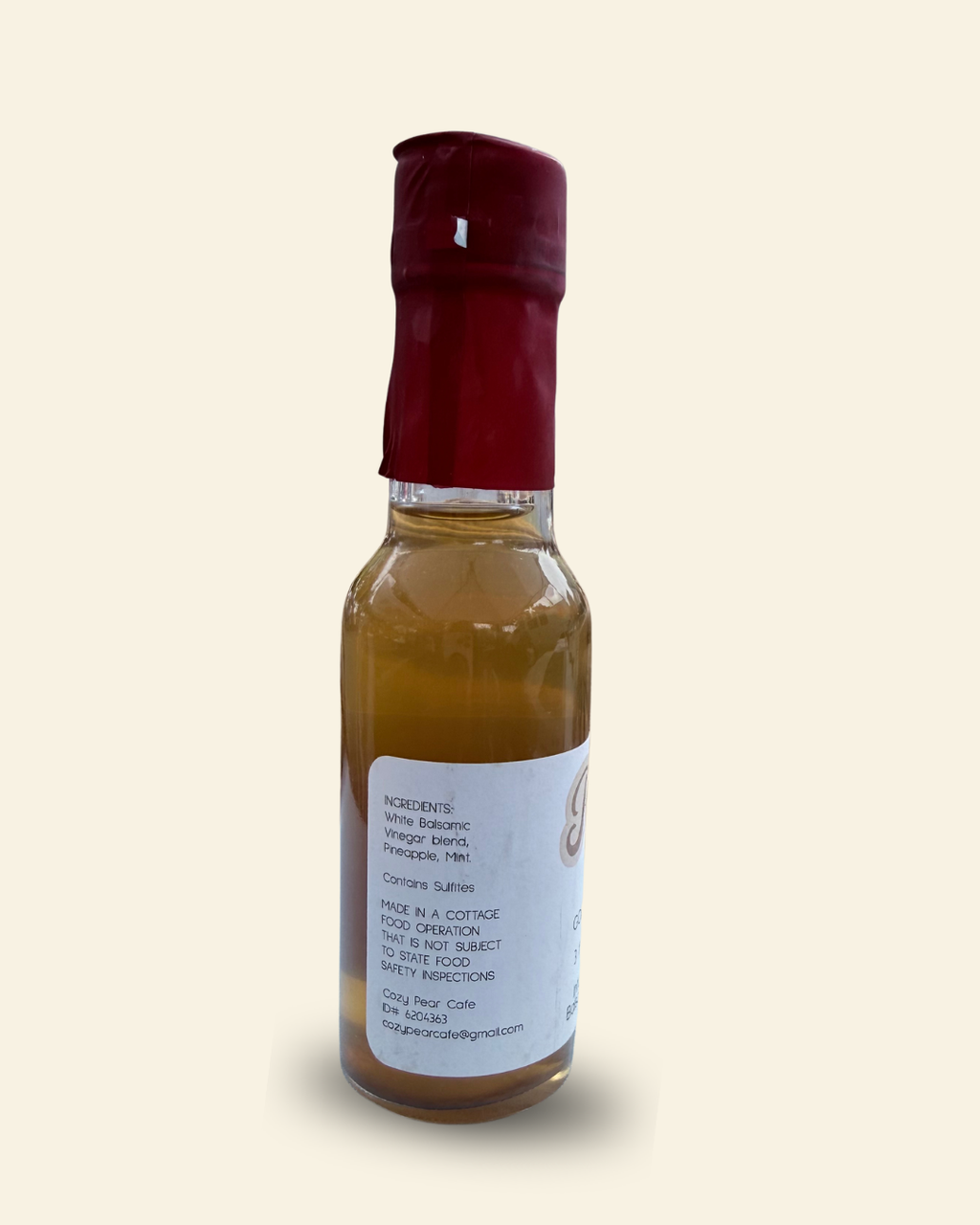 A small glass bottle of Pear Cafe Golden Breeze natural white vinegar with a dark red cap and a white label, set against a solid cream-colored background.