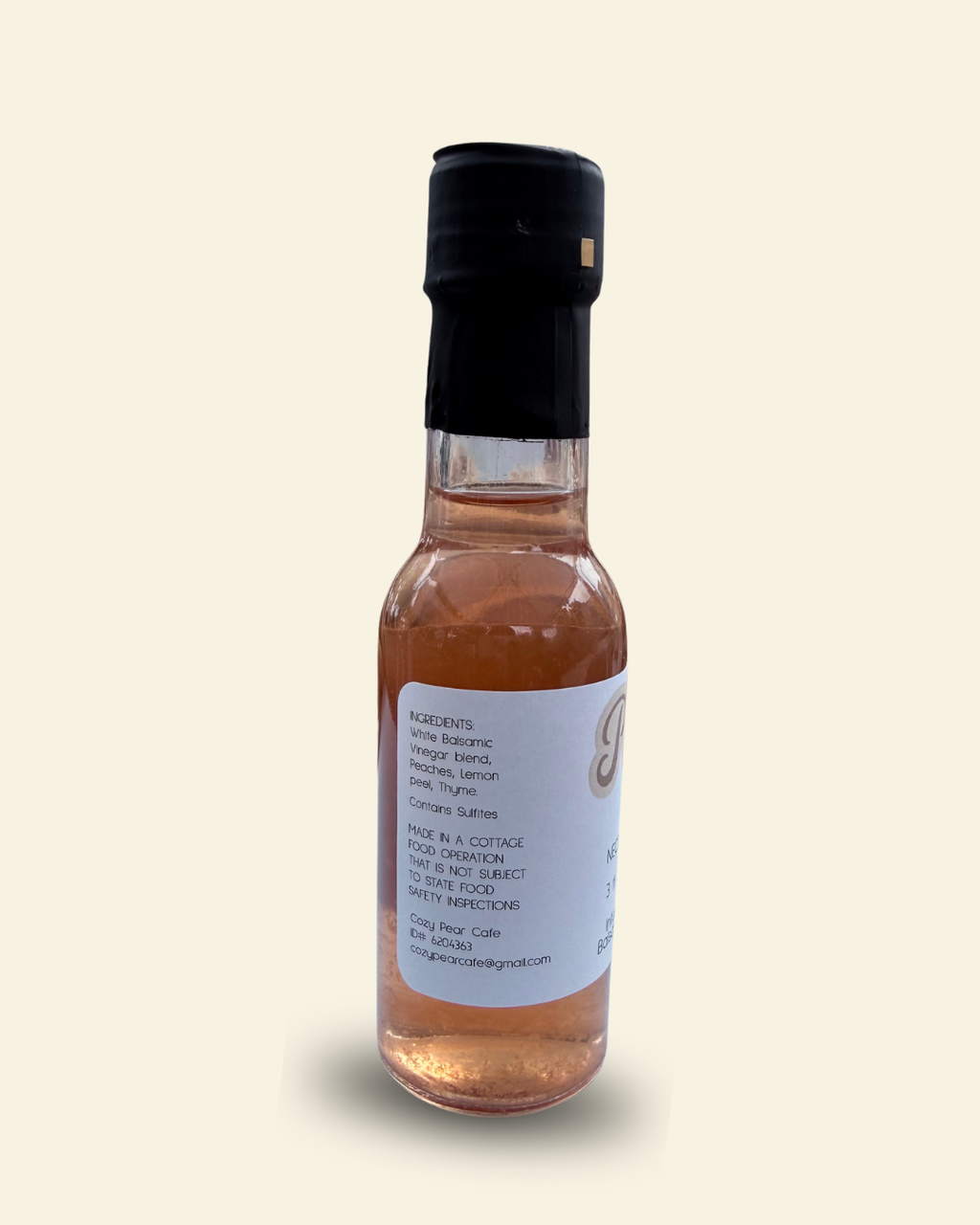 A small glass bottle of Cozy Pear Cafe Nectar & Vine infused white balsamic vinegar, featuring a white label and a black cap, set against a solid cream-colored background.