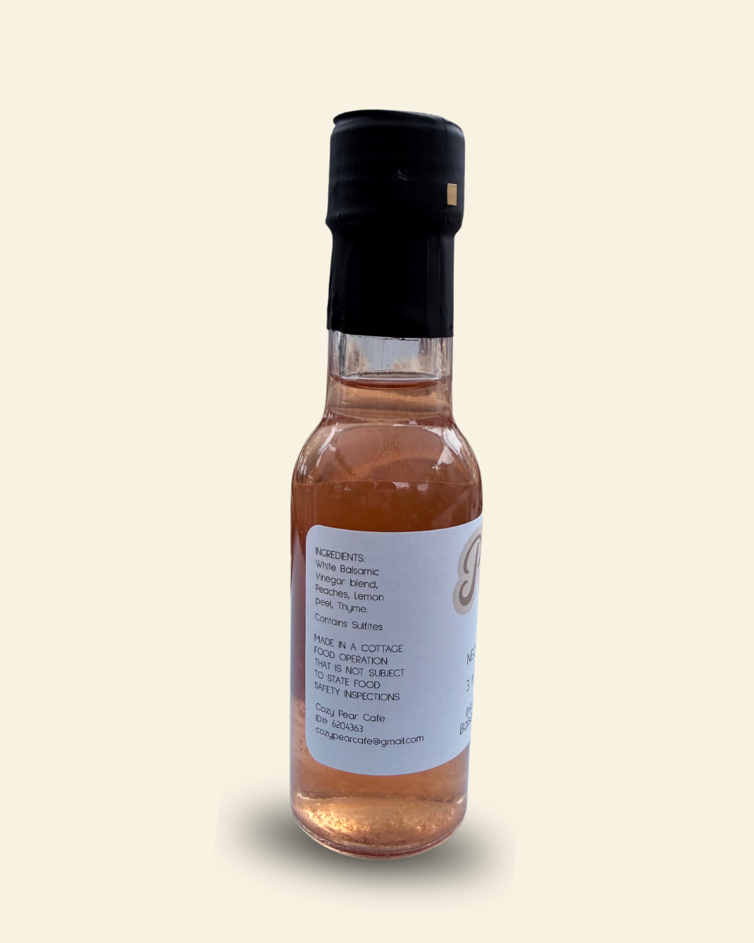 A small glass bottle of Cozy Pear Cafe Nectar & Vine infused white balsamic vinegar, featuring a white label and a black cap, set against a solid cream-colored background.