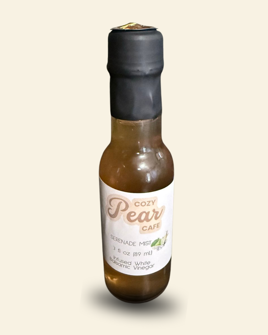 A small glass bottle of infused balsamic vinegar with amber-brown liquid, a black plastic cap, and a white label, shown against a plain light beige background.