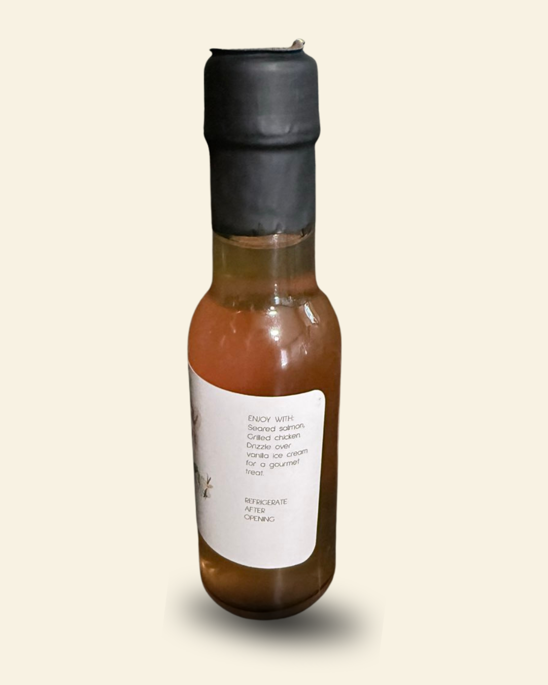 A small glass bottle of infused balsamic vinegar with amber-brown liquid, a black plastic cap, and a white label, shown against a plain light beige background.