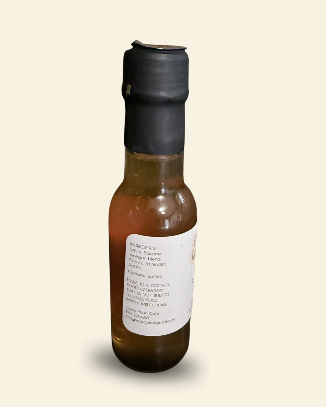 A small glass bottle of infused balsamic vinegar with amber-brown liquid, a black plastic cap, and a white label, shown against a plain light beige background.