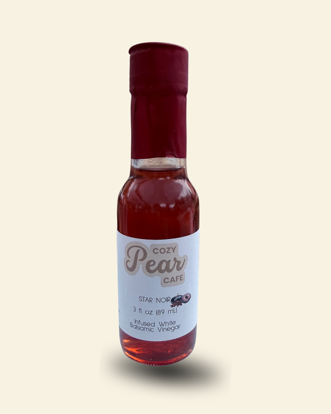 A small glass bottle of Cozy Pear Cafe Star Noir infused white balsamic vinegar, featuring a white label and a dark red cap, set against a solid pale cream background.