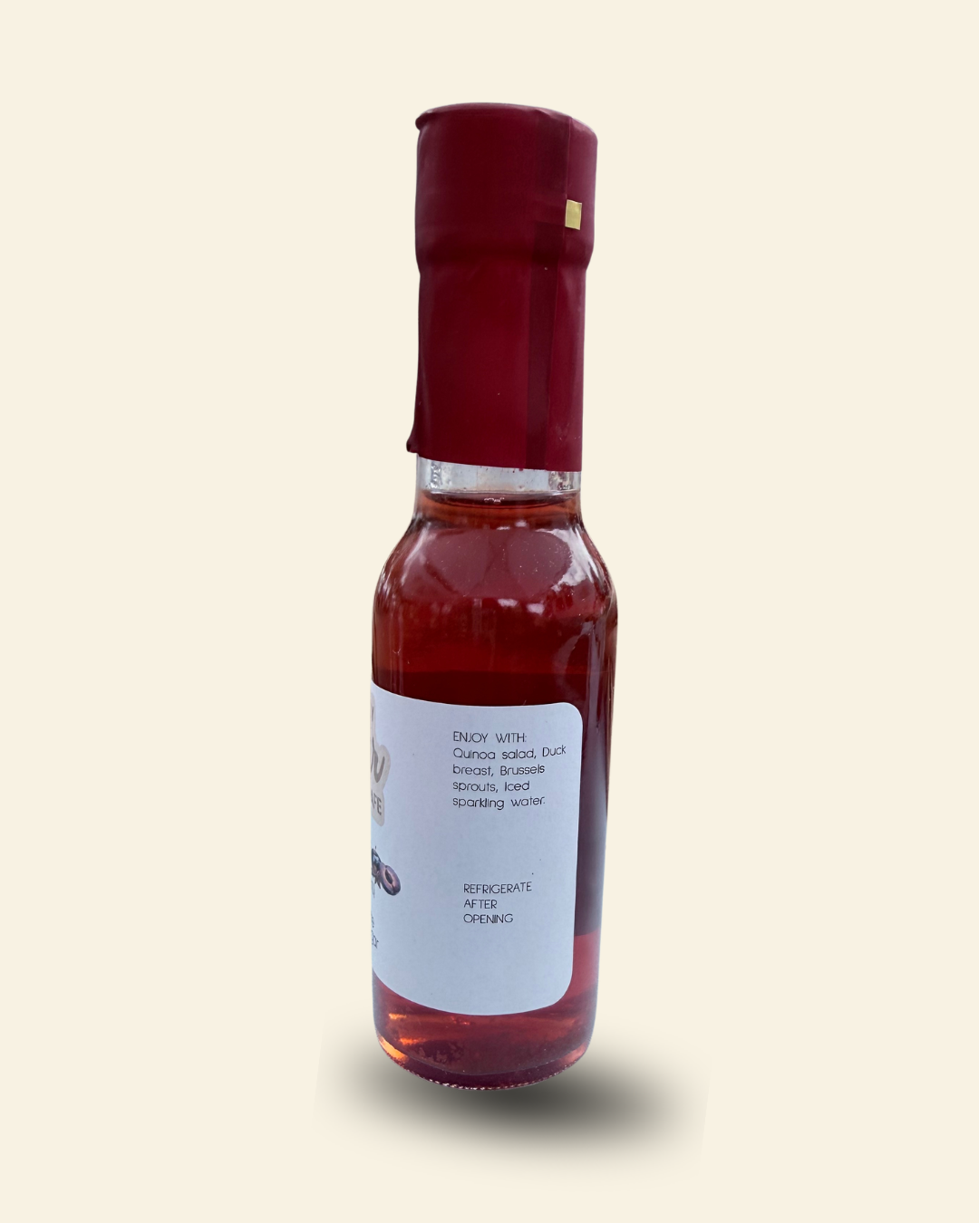 A small glass bottle of Cozy Pear Cafe Star Noir infused white balsamic vinegar, featuring a white label and a dark red cap, set against a solid pale cream background.