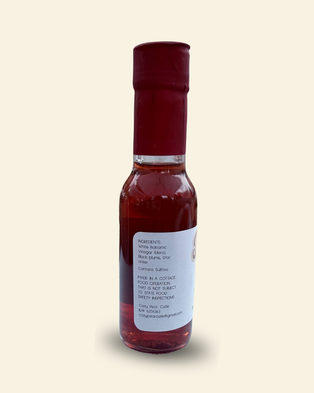 A small glass bottle of Cozy Pear Cafe Star Noir infused white balsamic vinegar, featuring a white label and a dark red cap, set against a solid pale cream background.