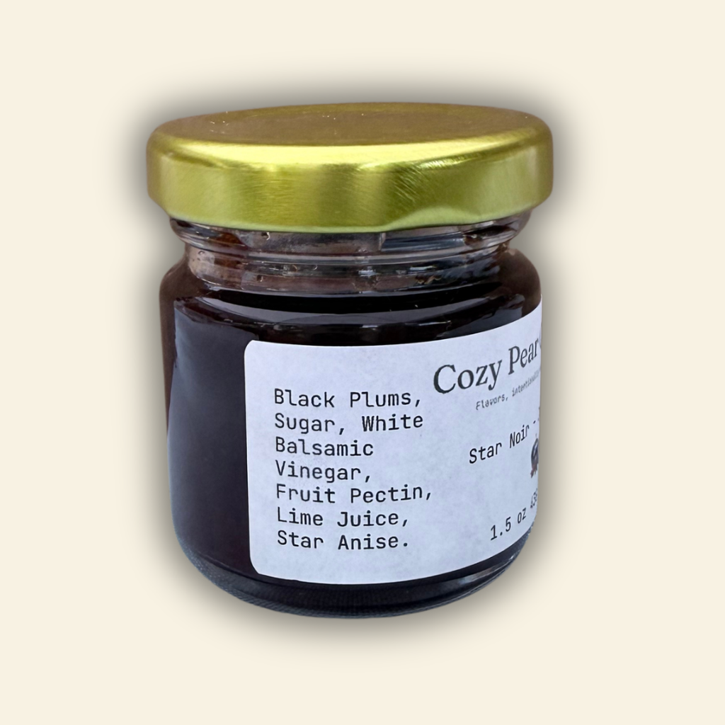 A jar of Cozy Pear Cafe Star Noir - Jam, featuring a white label and a metallic gold lid, set against a solid cream-colored background.