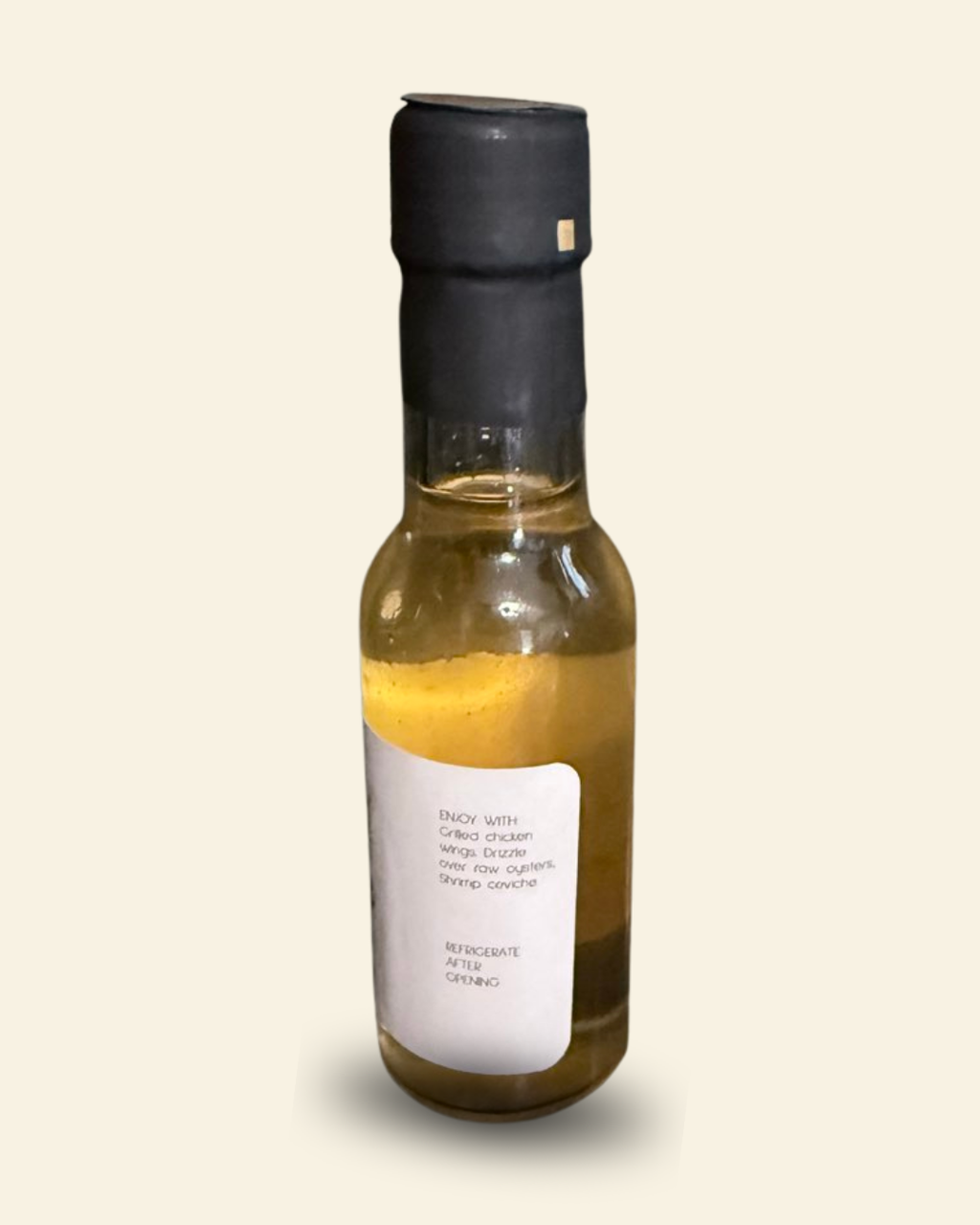 A small glass bottle of infused balsamic vinegar with a black cap and a white label, shown against a solid light beige background.