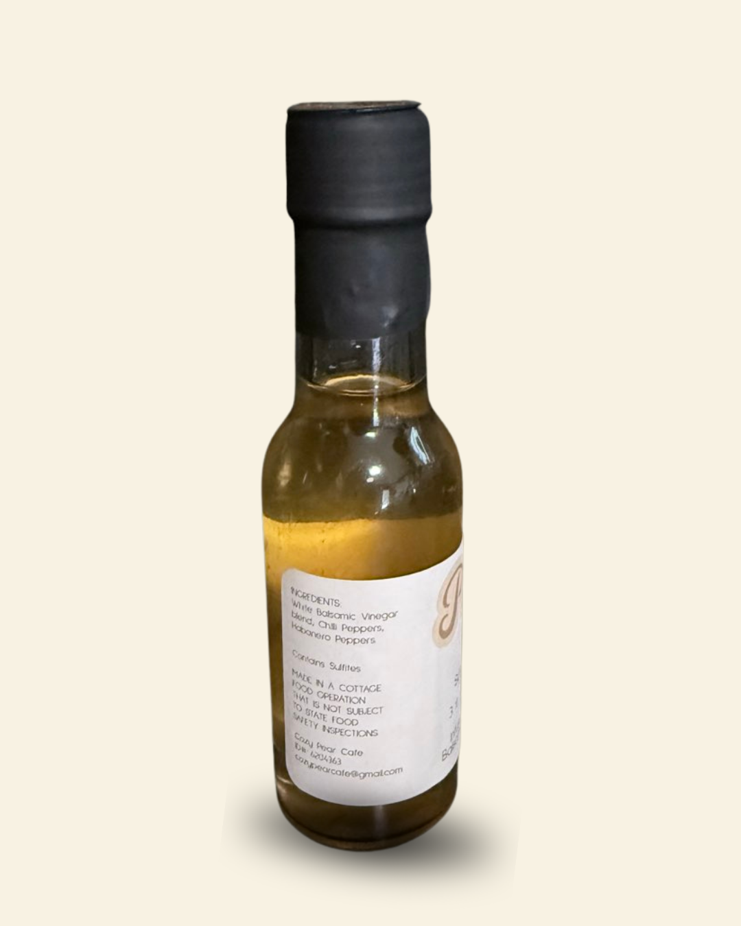 A small glass bottle of infused balsamic vinegar with a black cap and a white label, shown against a solid light beige background.