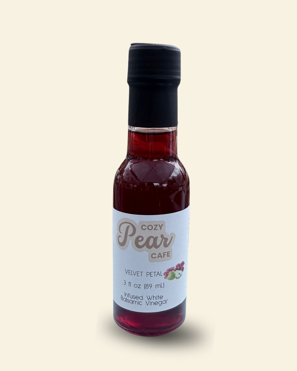 A small glass bottle of Cozy Pear Cafe Velvet Petal infused white balsamic vinegar, featuring a white label and a black cap, set against a solid pale cream background.