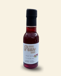A small glass bottle of Cozy Pear Cafe Velvet Petal infused white balsamic vinegar, featuring a white label and a black cap, set against a solid pale cream background.