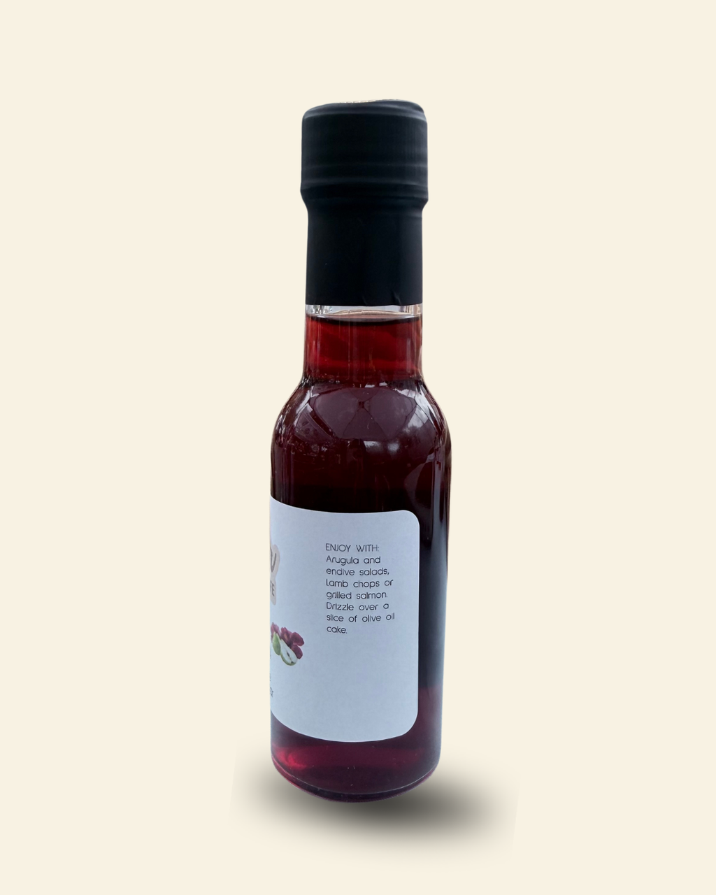 A small glass bottle of Cozy Pear Cafe Velvet Petal infused white balsamic vinegar, featuring a white label and a black cap, set against a solid pale cream background.