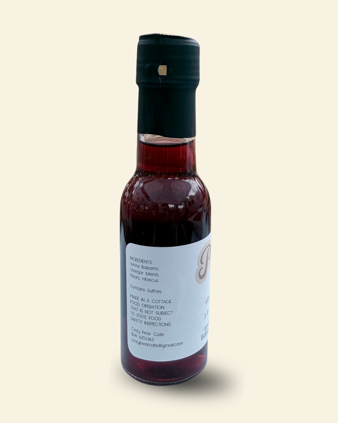 A small glass bottle of Cozy Pear Cafe Velvet Petal infused white balsamic vinegar, featuring a white label and a black cap, set against a solid pale cream background.